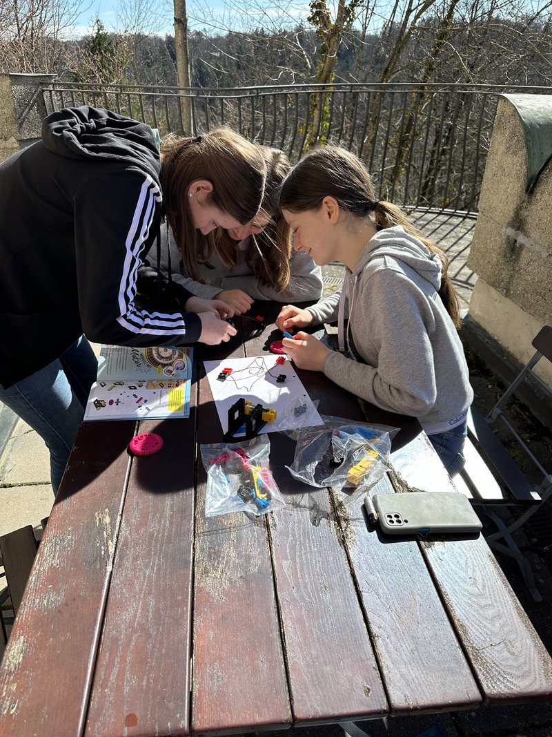 Das Bild zeigt drei Personen, die im Freien an einem Holztisch gemeinsam an einem kreativen Projekt arbeiten. Auf dem Tisch liegen bunte Materialien, Plastikbeutel mit Inhalt und ein Anleitungsblatt – eine handlungsorientierte Aktivität in naturnaher Umgebung.