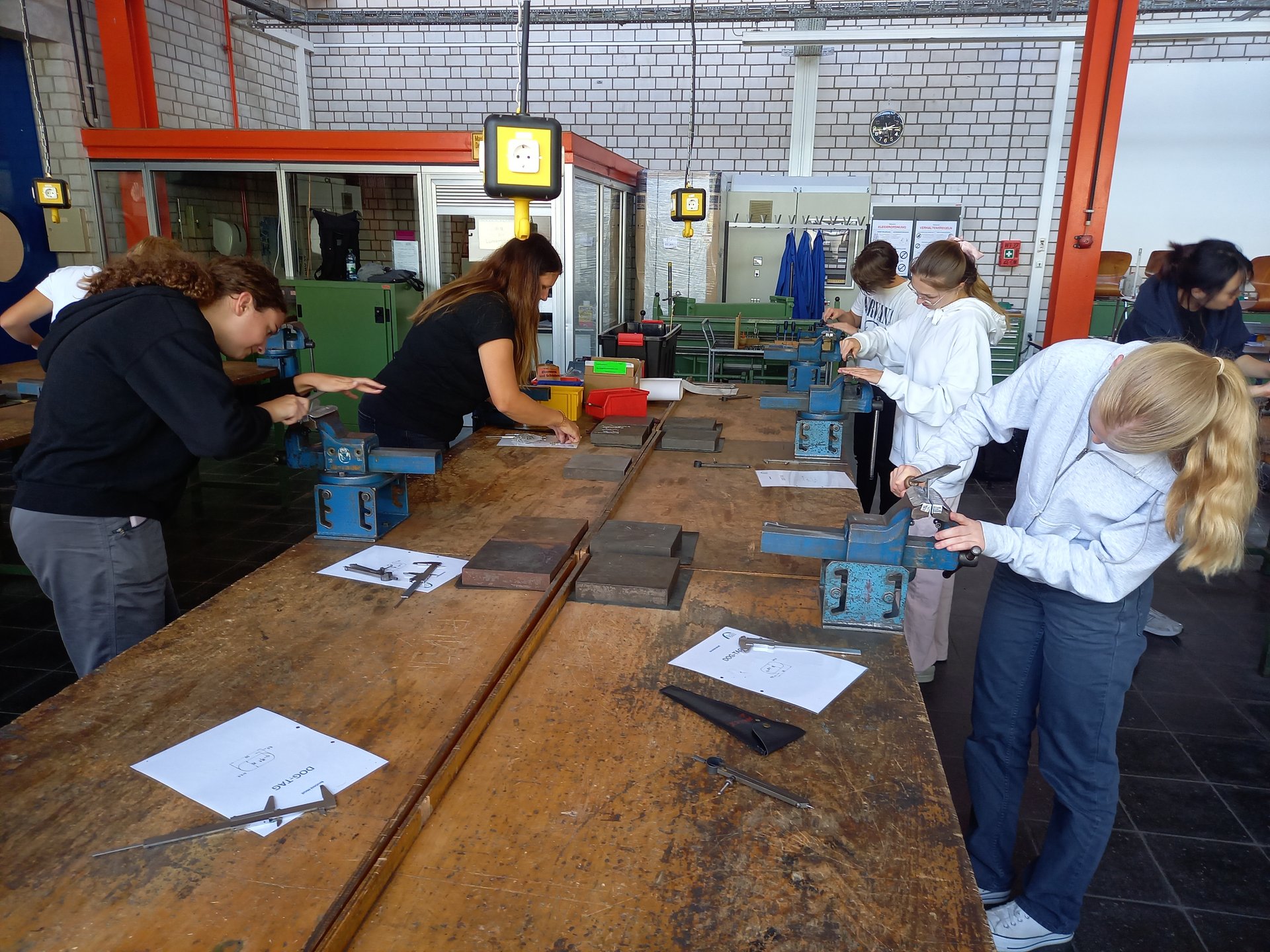 Das Bild zeigt mehrere Personen in einer Werkstatt, die an einem großen Holztisch mit Schraubstöcken und Werkzeugen arbeiten. Auf dem Tisch liegen Metallteile, Messinstrumente und Anleitungen – eine praxisorientierte Arbeitsumgebung mit Backsteinwänden und industrieller Beleuchtung.