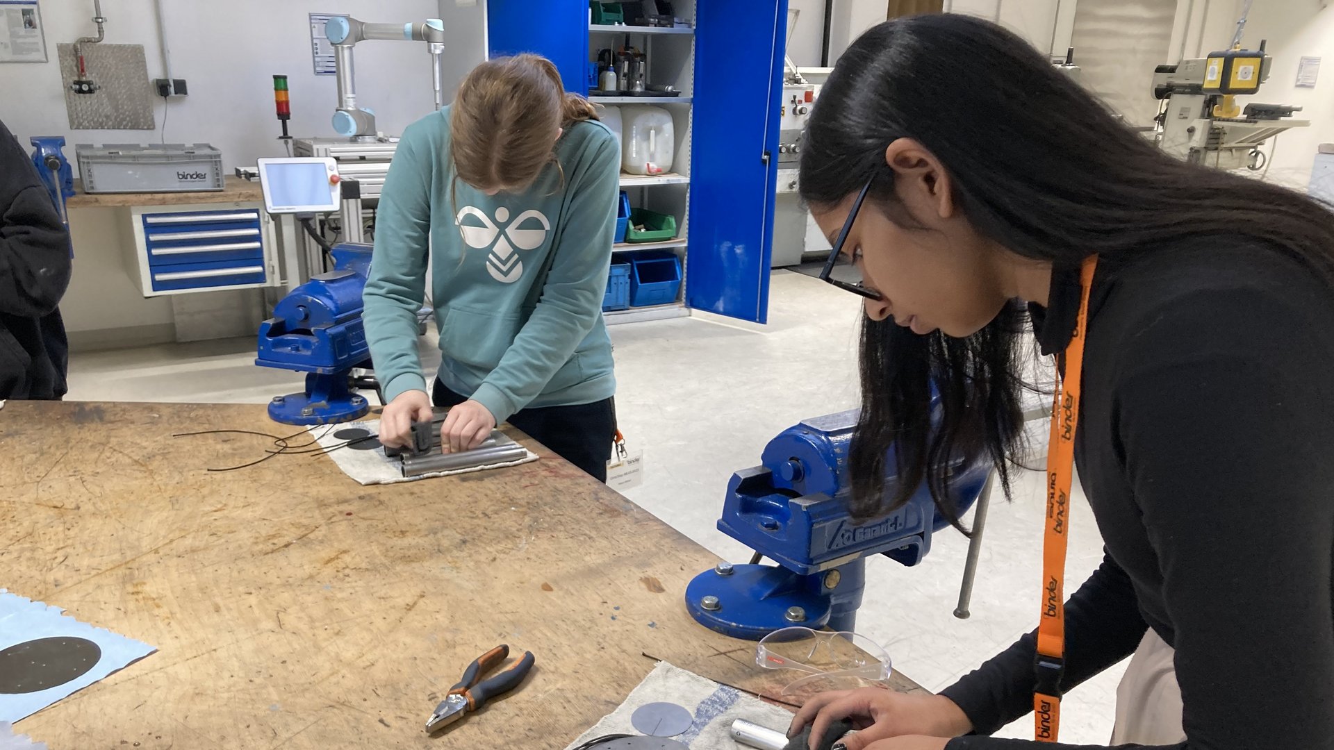 Das Bild zeigt zwei Personen in einer Werkstatt oder einem Labor, die mit Werkzeugen wie Zangen und Metallstäben arbeiten. Auf dem Tisch und im Hintergrund befinden sich weitere Geräte und Materialien – eine gut ausgestattete Umgebung für technische oder handwerkliche Tätigkeiten.