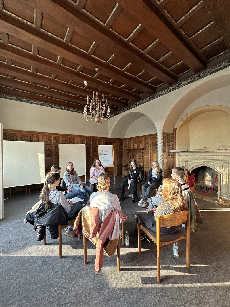 Das Bild zeigt eine Gruppe von Menschen, die in einem holzvertäfelten Raum mit Kamin, Kronleuchter und großen Fenstern im Kreis sitzt. Im Hintergrund stehen zwei Whiteboards und ein Flipchart – eine ruhige, lichtdurchflutete Umgebung für gemeinsames Arbeiten oder Austausch.