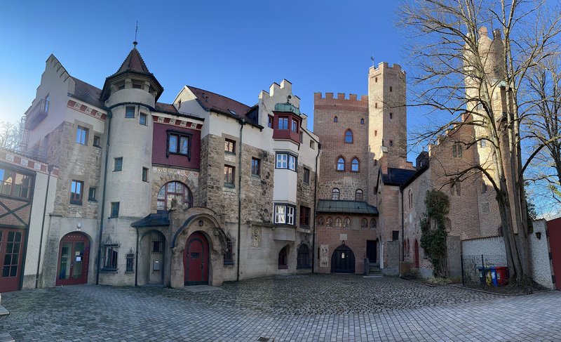 Das Bild zeigt ein historisches, burgähnliches Gebäude mit mehreren Türmen und dekorativen Elementen aus Stein und Backstein. Vor dem Gebäude liegt ein gepflasterter Innenhof, rechts steht ein großer Baum – alles bei klarem, blauem Himmel.