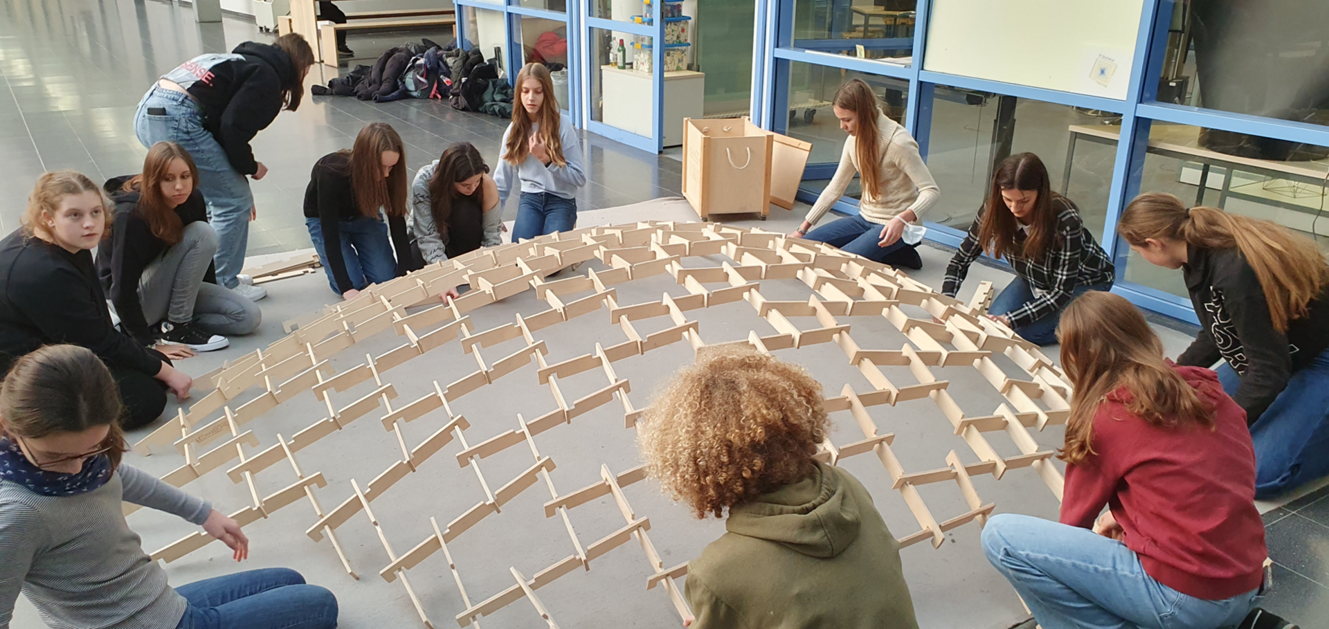 Das Bild zeigt mehrere Personen, die gemeinsam eine große, kuppelförmige Struktur aus ineinandergreifenden Holzteilen aufbauen. Sie sitzen oder knien um das entstehende Gerüst in einem hellen Innenraum mit großen Fenstern – eine kreative Gruppenarbeit mit handwerklichem Fokus.