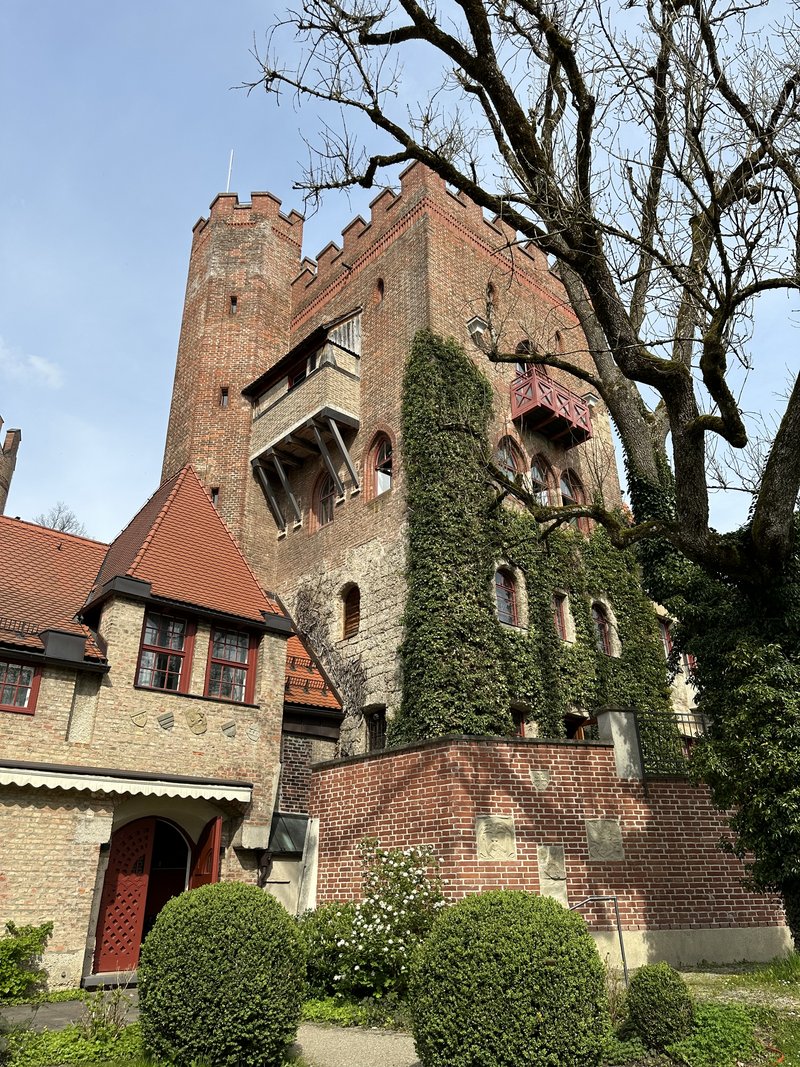 Das Bild zeigt ein historisches Backsteinschloss mit einem hohen Turm, bewachsen mit Efeu und umgeben von gepflegten Büschen – ein eindrucksvolles Beispiel denkmalgeschützter Architektur bei klarem Tageslicht.