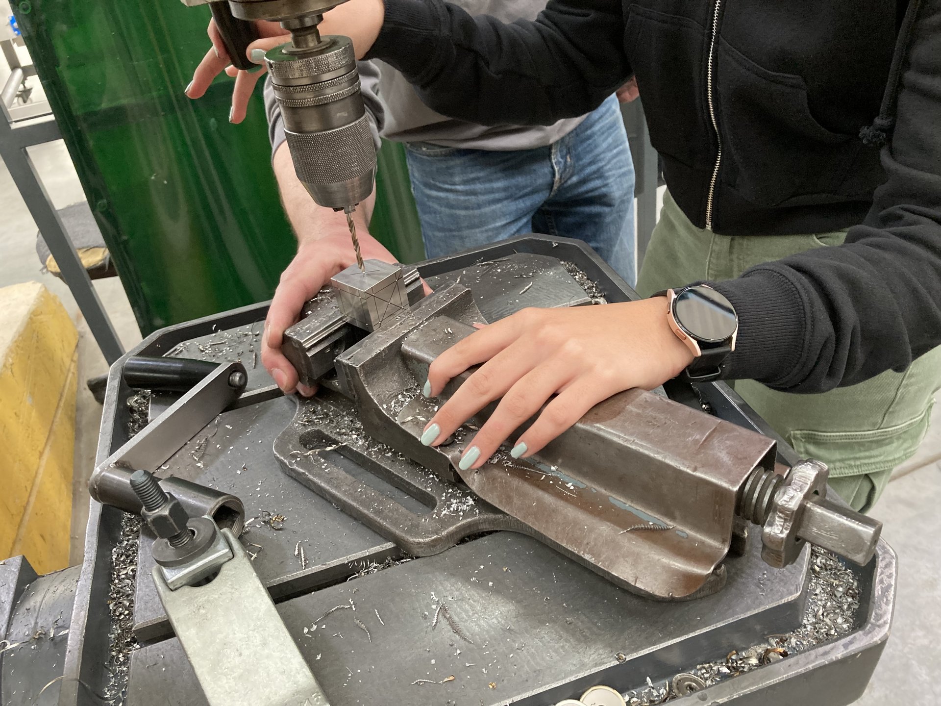 Das Bild zeigt zwei Personen in einer Werkstatt, die gemeinsam an einer Ständerbohrmaschine arbeiten. Eine Person bedient die Bohrmaschine, während die andere ein markiertes Metallstück in einem Schraubstock fixiert – Metallspäne auf dem Tisch deuten auf aktive Bohrarbeiten hin.