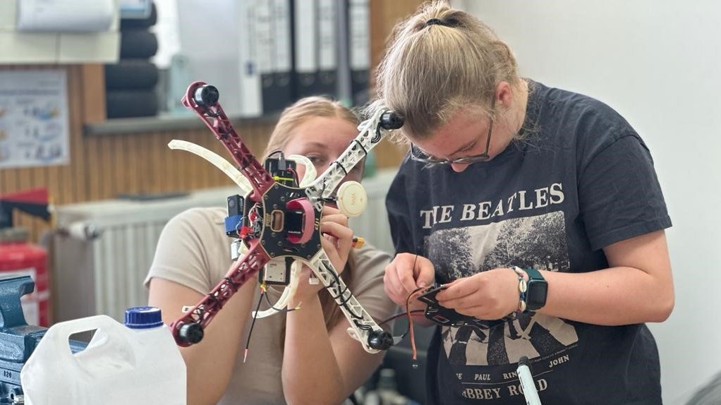 Das Bild zeigt zwei Personen in einer Werkstatt oder einem Labor, die gemeinsam an einem Drohnenmodell arbeiten. Eine Person hält den Rahmen, während die andere Kabel anschließt. Auf dem Tisch liegen Werkzeuge, technische Geräte und ein großer Behälter mit Flüssigkeit.