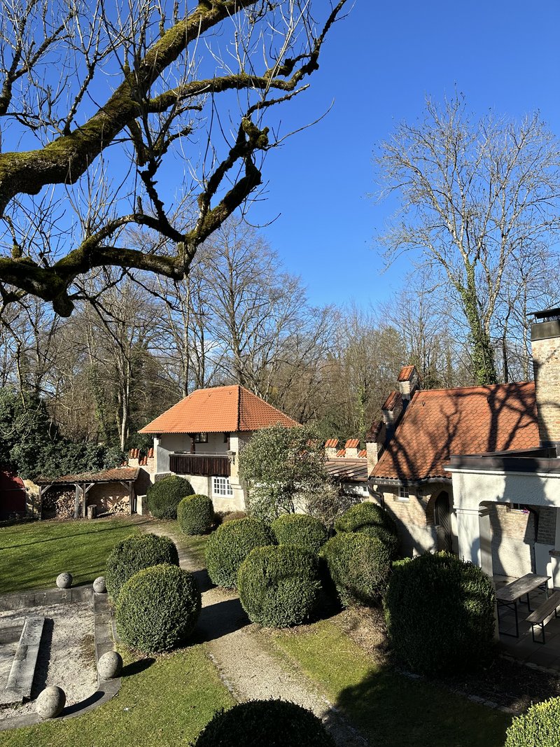 Das Bild zeigt einen gepflegten Garten mit Kieswegen und akkurat geschnittenen Büschen. Zwei Gebäude mit roten Ziegeldächern stehen im Vorder- und Hintergrund, umgeben von hohen, blattlosen Bäumen – ein ruhiger Außenbereich bei klarem, blauem Himmel.