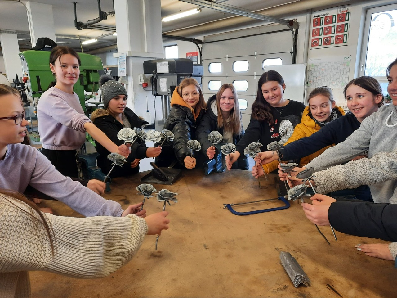 Mehrere Personen stehen um einen Holztisch in einer Werkstattumgebung, jede hält ein kleines metallisches Objekt in der Hand. Im Hintergrund sind Werkzeuge und Geräte zu sehen.