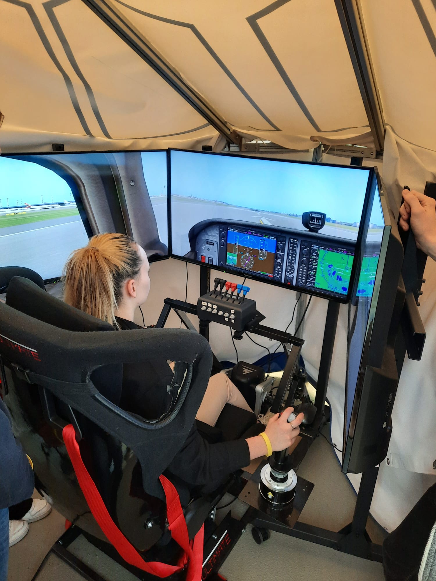 Das Bild zeigt eine Person in einem Flugsimulator, sitzend in einem Rennsitz mit roten Gurten und umgeben von mehreren Monitoren, die ein Cockpit mit Fluginstrumenten darstellen. Vor der Person befinden sich Steuerknüppel, Schalter und Bedienfelder – eine realitätsnahe Trainingsumgebung für Flugerfahrungen.