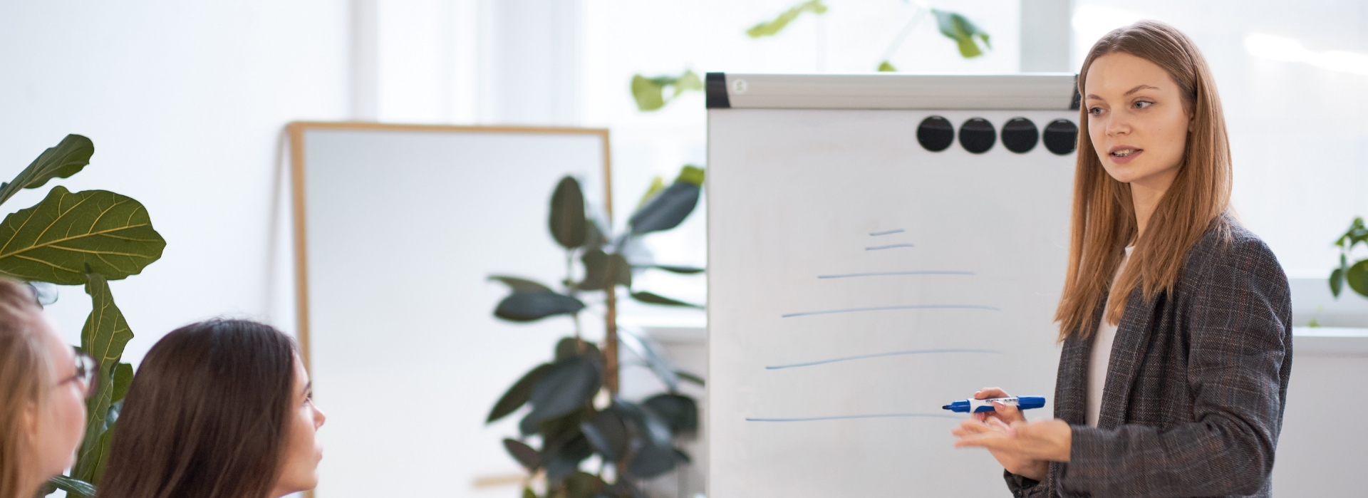 Eine Person steht vor einem Flipchart und hält eine Präsentation oder einen Vortrag. Im Hintergrund sind weitere Personen und Pflanzen zu sehen, der Text auf dem Flipchart ist nicht lesbar.