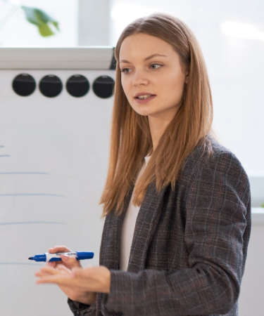 Eine Person steht vor einem Flipchart und hält eine Präsentation oder einen Vortrag. Im Hintergrund sind weitere Personen und Pflanzen zu sehen, der Text auf dem Flipchart ist nicht lesbar.