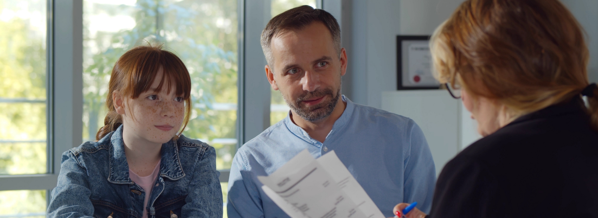 Ein Mann und ein junges Mädchen sitzen gemeinsam an einem Tisch und sprechen mit einer Person, die ihnen Dokumente zeigt – vermutlich in einem Beratungsgespräch. Im Hintergrund ist durch ein Fenster Tageslicht zu sehen.