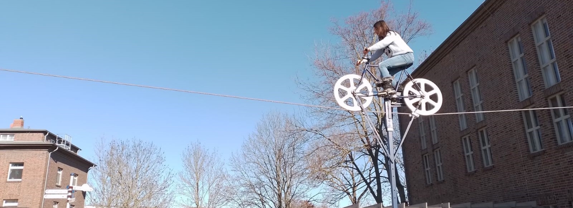 Eine Person fährt mit einem Fahrrad auf einem Seil in der Luft, im Hintergrund sind Gebäude, Bäume und ein klarer blauer Himmel zu sehen.