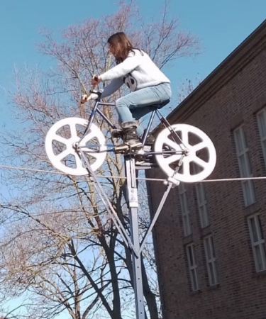 Eine Person fährt mit einem Fahrrad auf einem Seil in der Luft, im Hintergrund sind Gebäude, Bäume und ein klarer blauer Himmel zu sehen.