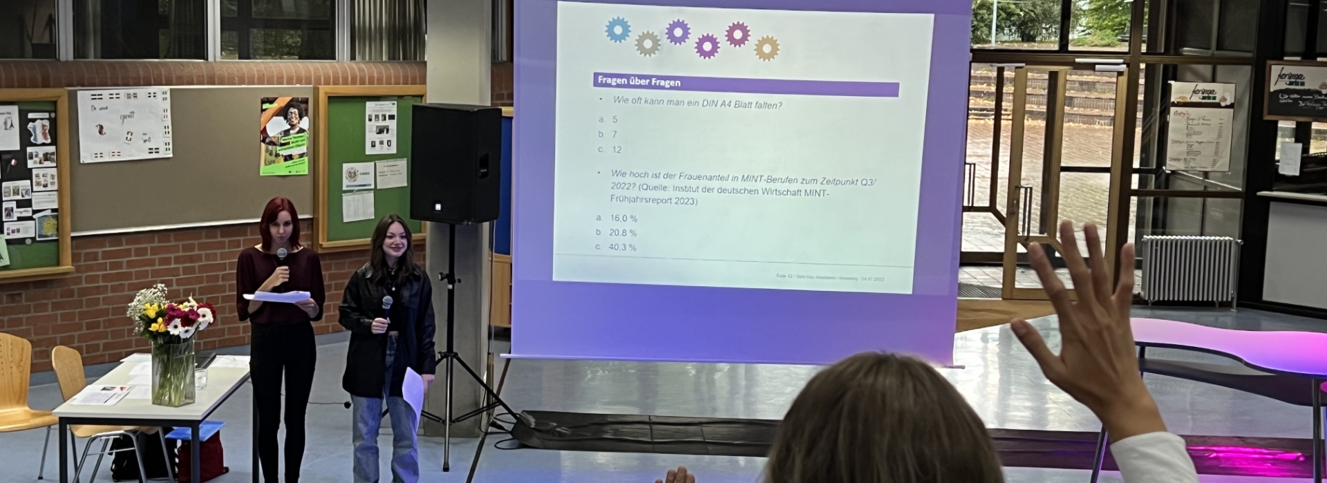 In einem Raum mit mehreren Personen findet eine Präsentation statt. Auf einem großen Bildschirm ist eine Folie mit mathematischen Funktionen und Gleichungen zu sehen, eine Person steht vorne und präsentiert.