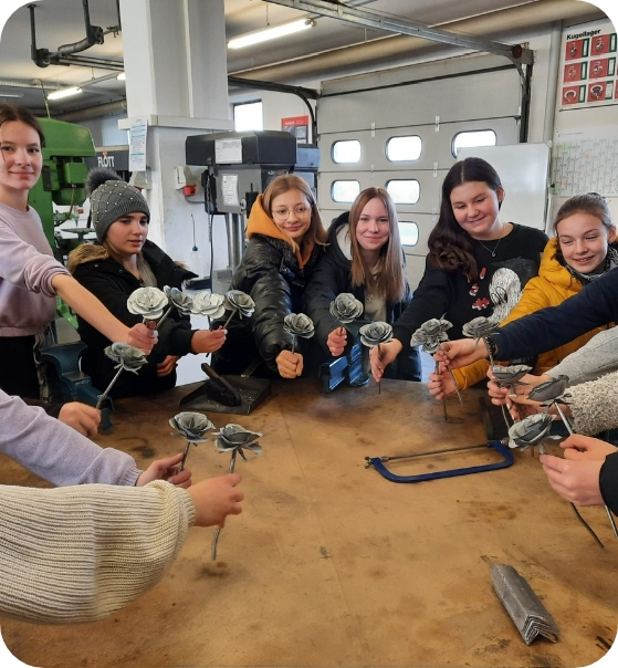Mehrere Personen stehen um einen Holztisch in einer Werkstattumgebung, jede hält ein kleines metallisches Objekt in der Hand. Im Hintergrund sind Werkzeuge und Geräte zu sehen.