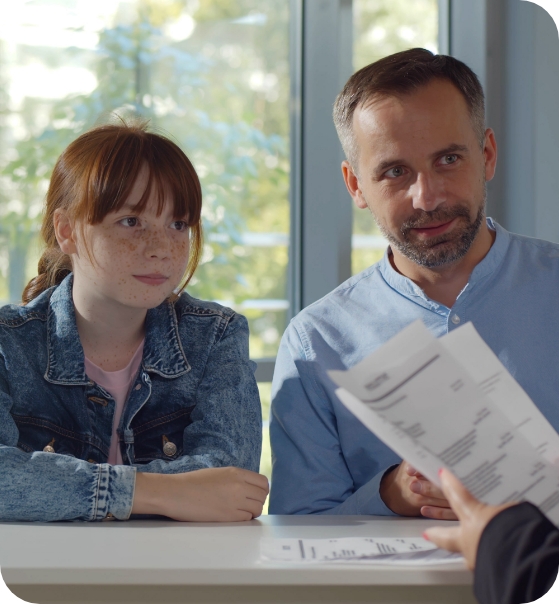 Ein Mann und ein junges Mädchen sitzen gemeinsam an einem Tisch und sprechen mit einer Person, die ihnen Dokumente zeigt – vermutlich in einem Beratungsgespräch. Im Hintergrund ist durch ein Fenster Tageslicht zu sehen.