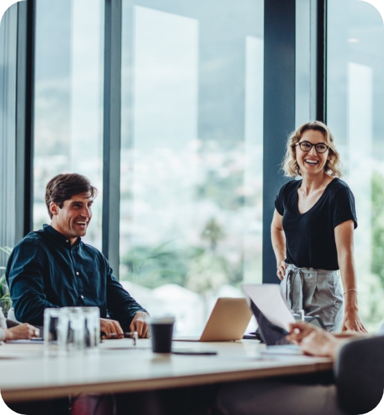Drei Personen befinden sich in einem modernen Büro. Eine Person steht, zwei sitzen an einem Tisch mit Papieren, einem Laptop und Tassen. Große Fenster im Hintergrund lassen Tageslicht herein und geben den Blick auf eine städtische Umgebung frei.